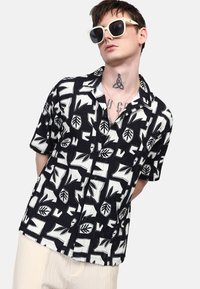 Camicia a maniche corte con bottoni, caratterizzata da un motivo a foglie in bianco e nero. Tessuto testurizzato con vestibilità rilassata e colletto.