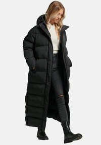 Sort polstret lang frakke med høj krave, der har en knaplukning og strukturerede sektioner. Bæres med en cremefarvet sweater og sorte jeans.