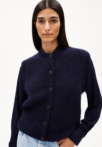 Marineblauer Strickweste mit rundem Ausschnitt, Frontknopfverschluss und strukturiertem Muster. Kombiniert mit einem dunklen Langarmshirt darunter.