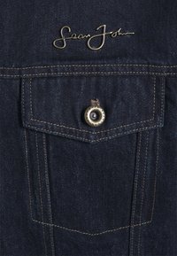 Chemise en denim foncé avec une poche poitrine cousue, dotée d'un bouton-pression doré, et d'un logo en cursive au-dessus de la poche en fils métalliques.