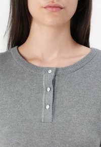 Femme portant un haut gris en maille avec un col rond et trois petits boutons blancs sur la patte de boutonnage devant, vue des épaules au menton.