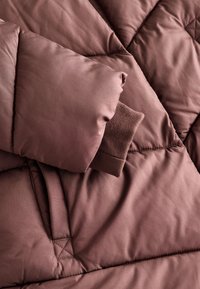 Brązowa kurtka puchowa z pikowaniem w kształcie diamentu, o pluszowej fakturze. Manżety wykonane z ciemniejszej brązowej dzianiny.