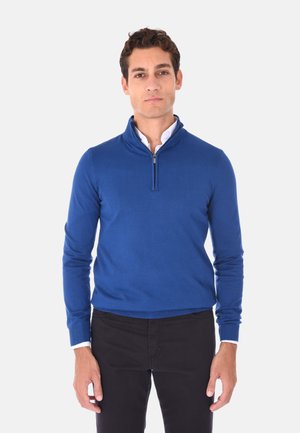 Maglione blu con colletto a mezza zip, maniche lunghe e texture liscia, abbinato a una camicia bianca con colletto sotto.