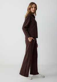 Abbigliamento marrone, testurizzato, composto da una maglia ampia con colletto alto e pantaloni a gamba larga, abbinati a scarpe da ginnastica bianche.