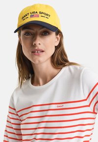 Gul kasket med marineblå skygge, der har "GANT USA SPORT" og et lille flaglogo. Hvid skjorte med røde vandrette striber og "GANT" logo.