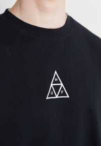 Black sweatshirt with a round neckline featuring a white embroidered triangular logo with the letters H, U, and F. Soft fabric texture.