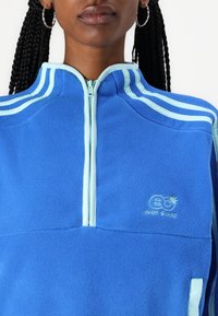Maglia in pile azzurra con mezza zip, rifiniture in azzurro chiaro e logo ricamato. Presenta un colletto alto e un tessuto testurizzato.