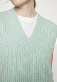 Lichtgroene vest met een diepe V-hals, gebreide textuur en brede armsgaten, gedragen over een wit T-shirt. Zacht, comfortabel materiaal.