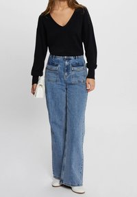 Mujer con blusa negra de cuello en V y manga larga, vaqueros anchos de tiro alto y color azul con bolsillos frontales, botas blancas y sosteniendo un bolso blanco.