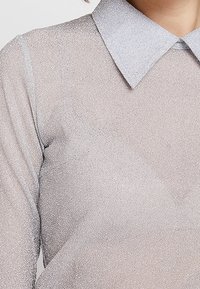 Haut argenté scintillant avec un col en forme de col chemise, présentant un tissu texturé métallique et des manches longues.