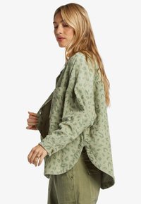 Mujer con cabello rubio largo, vestida con una camisa de pana de leopardo verde claro y pantalones cargo verde oliva, posando de lado sobre un fondo blanco.