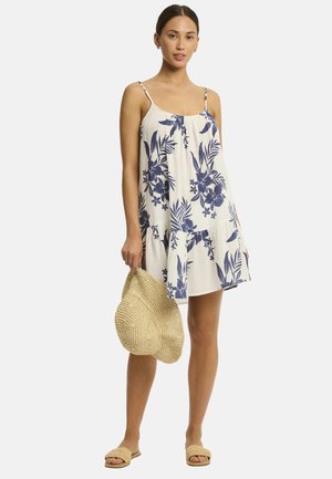 Eine junge Frau steht in einem blumengemusterten Sommerkleid und hält eine Strohtasche. Sie hat einen neutralen Gesichtsausdruck und steht barfuß vor einem schlichten Hintergrund.
