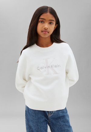 Dievča s dlhými tmavými vlasmi, oblečené v bielom svetri Calvin Klein a modrých džínsoch, stojace pred jednoduchým sivým pozadím.