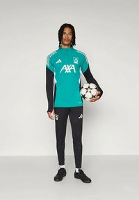 Zaļš un melns futbola treniņtērps ar garām piedurknēm, ar logo un baltiem akcentiem, kombinēts ar melnām biksēm un futbola bumbu.