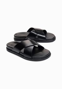 Paire de sandales slides noires avec larges brides croisées et semelles plates sur fond blanc.