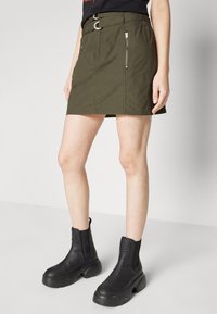 Olíva zöld farmer mini szoknya két cipzáros zsebével és fémgyűrűs övével. Fekete, vaskos bokacsizmával párosítva.