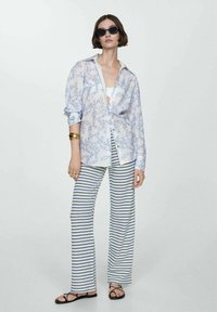 Camicia leggera a maniche lunghe con stampa floreale bianca e blu, abbinata a pantaloni a gamba larga a righe blu e bianchi. Sandali neri e occhiali da sole.