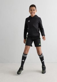 Svart hettegenser med frontlomme, svarte shorts med grønne detaljer, svarte fotballsokker med hvite striper, og svarte fotballsko med grønn dekor. 