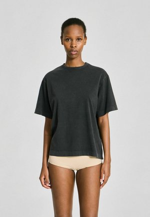Femme debout, face à l'avant, portant un t-shirt noir ample et des sous-vêtements beiges, sur un fond uni gris clair.