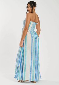 Halterlös maxiklänning med smockad bodice, med blå och gröna geometriska mönster på en gräddvit bakgrund. Lång, flödande silhuett.