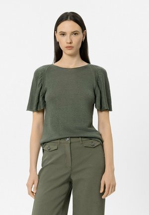 Junge Frau mit langen dunklen Haaren, die einen olivgrünen kurzärmligen Pullover und passende Hose trägt, vor einem schlichten weißen Hintergrund stehend.