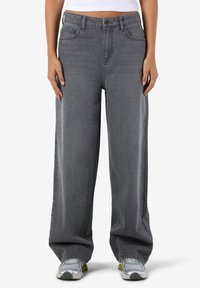 Henkilö, jolla on päällään väljät harmaat farkut ja hopeiset lenkkitossut, seisoo valkoista taustaa vasten kädet rentona sivuilla.