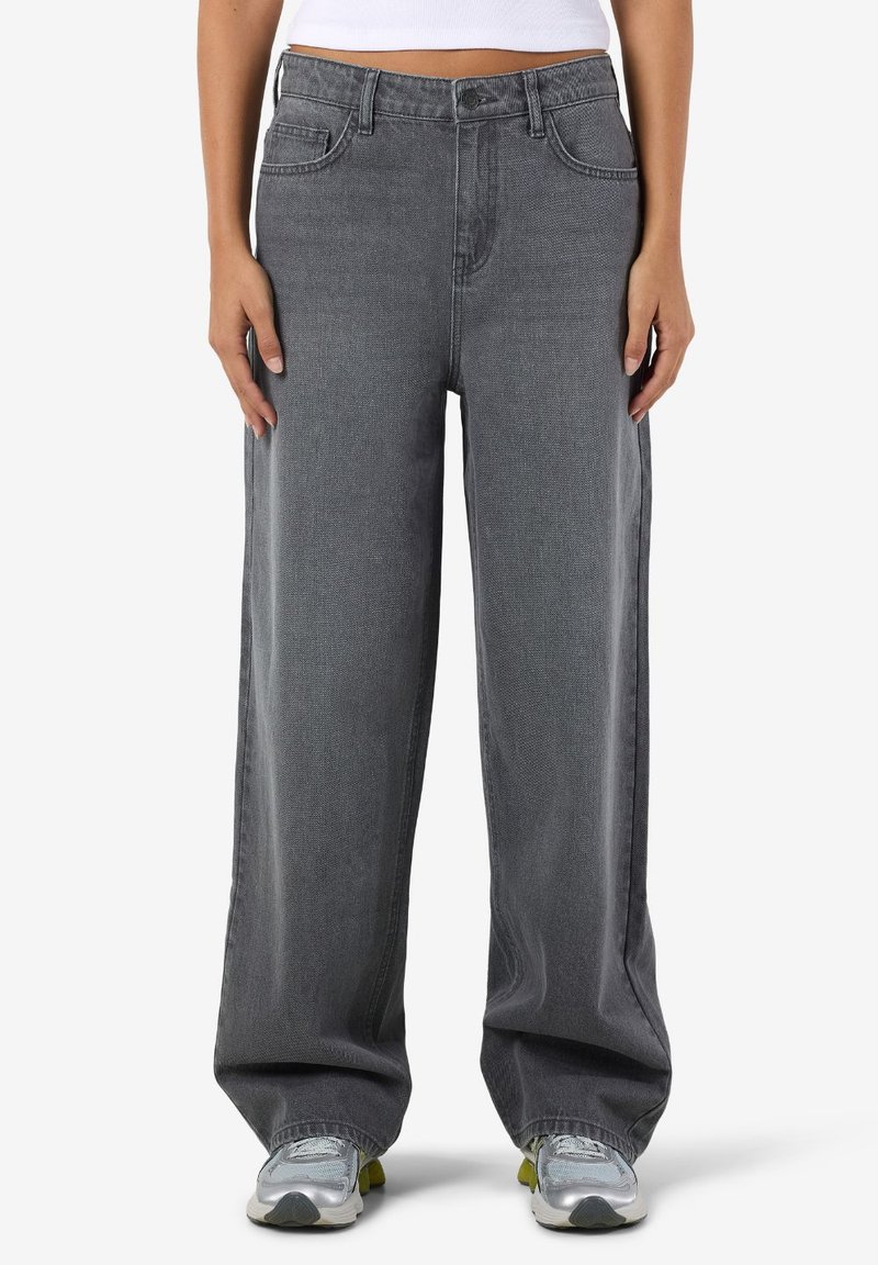 Henkilö, jolla on p äällään väljät harmaat farkut ja hopeiset lenkkitossut, seisoo valkoista taustaa vasten kädet rentona sivuilla.