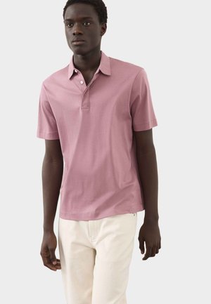 Ung mand iført en lyserød polo med korte ærmer og knapper samt lyse bukser, stående foran en ensfarvet lys baggrund.