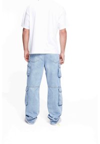 Man med ljusblå denim cargo-byxor och en enfärgad vit t-shirt med korta ärmar, står med ryggen mot kameran.