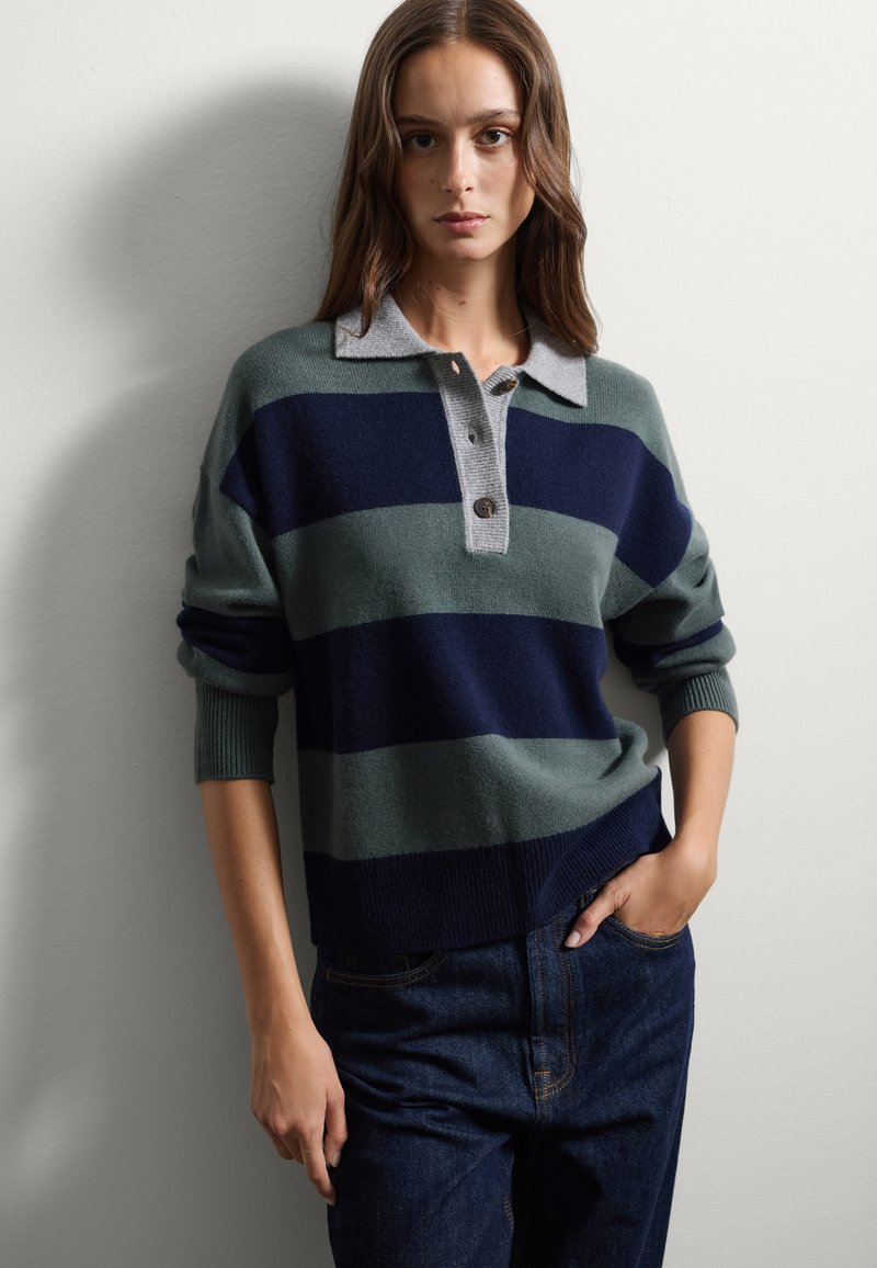 Suéter a rayas en verde y marino con un cuello gris. Presenta una tapeta de botones, puños acanalados y un ajuste relajado, combinado con pantalones oscuros.