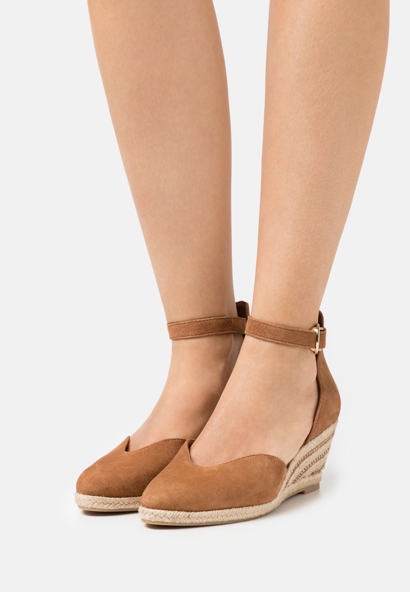 Chaussures compensées en daim marron avec brides chevilles et semelles en corde tressée, portées pieds nus sur un fond clair uni.