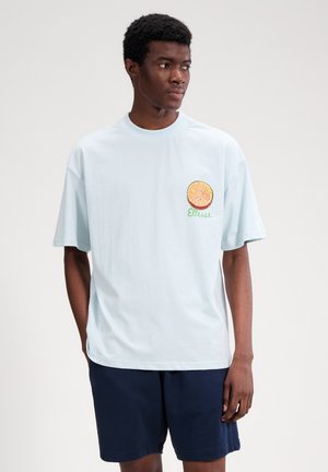 Mladi moški, oblečen v svetlomodro majico s kratkimi rokavi z logotipom citrusnega sadja in napisom »Ellesse«, v kombinaciji s temnomodrimi kratkimi hlačami, stoji na enostavni podlagi.