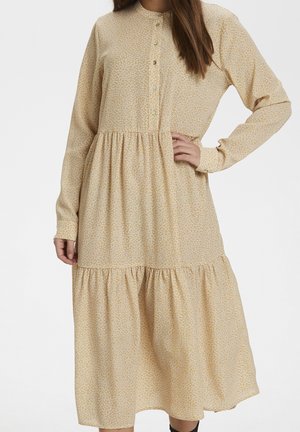 Femme portant une robe midi à manches longues, beige clair à imprimé floral, avec des boutons sur le haut devant et des volants froncés à la jupe.
