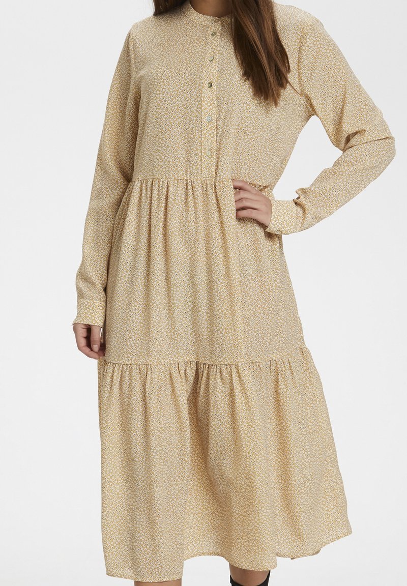 Femme portant une robe midi à manches longues, beige clair à imprimé floral, avec des boutons sur le haut devant et des volants froncés à la jupe.