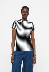 Γυναίκα όρθια, φορώντας κοντομάνικο πουκάμισο με μαύρες και άσπρες ρίγες και φαρδιά μπλε παντελόνια, σε λευκό και απλό φόντο.