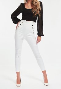 Sort gennemsigtig bluse med knapper foran og lange ærmer, parret med højhalsede hvide bukser med guldkneppe-detaljer.