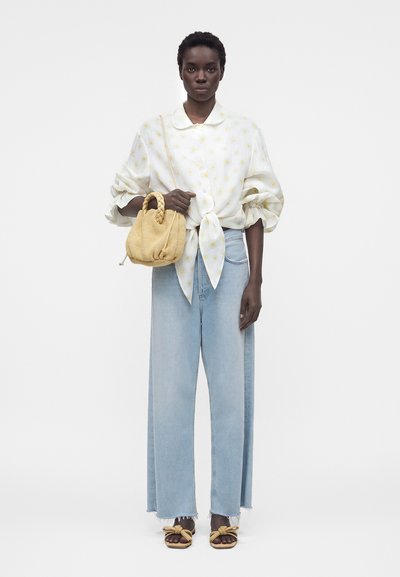 Γυναίκα που φοράει λευκό μπλουζάκι δεμένο στη μέση με κίτρινο λουλουδάτο μοτίβο, ανοιχτό μπλε τζιν παντελόνι με φαρδιά πόδια, κίτρινη πλεκτή τσάντα και αντίστοιχα σανδάλια.