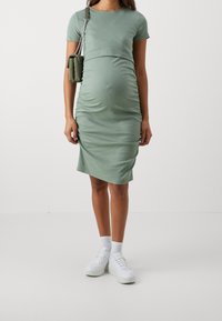 Grünes, tailliertes Midikleid aus weichem Stoff, mit kurzen Ärmeln und seitlichem Raffung; kombiniert mit weißen Sneakers und einer kleinen grünen Handtasche.