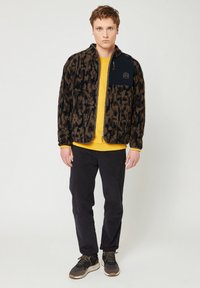Giacca in pile a motivo marrone e nero, con chiusura a cerniera e tasca sul petto nera. Indossata sopra una maglietta gialla e abbinata a pantaloni scuri e sneakers.