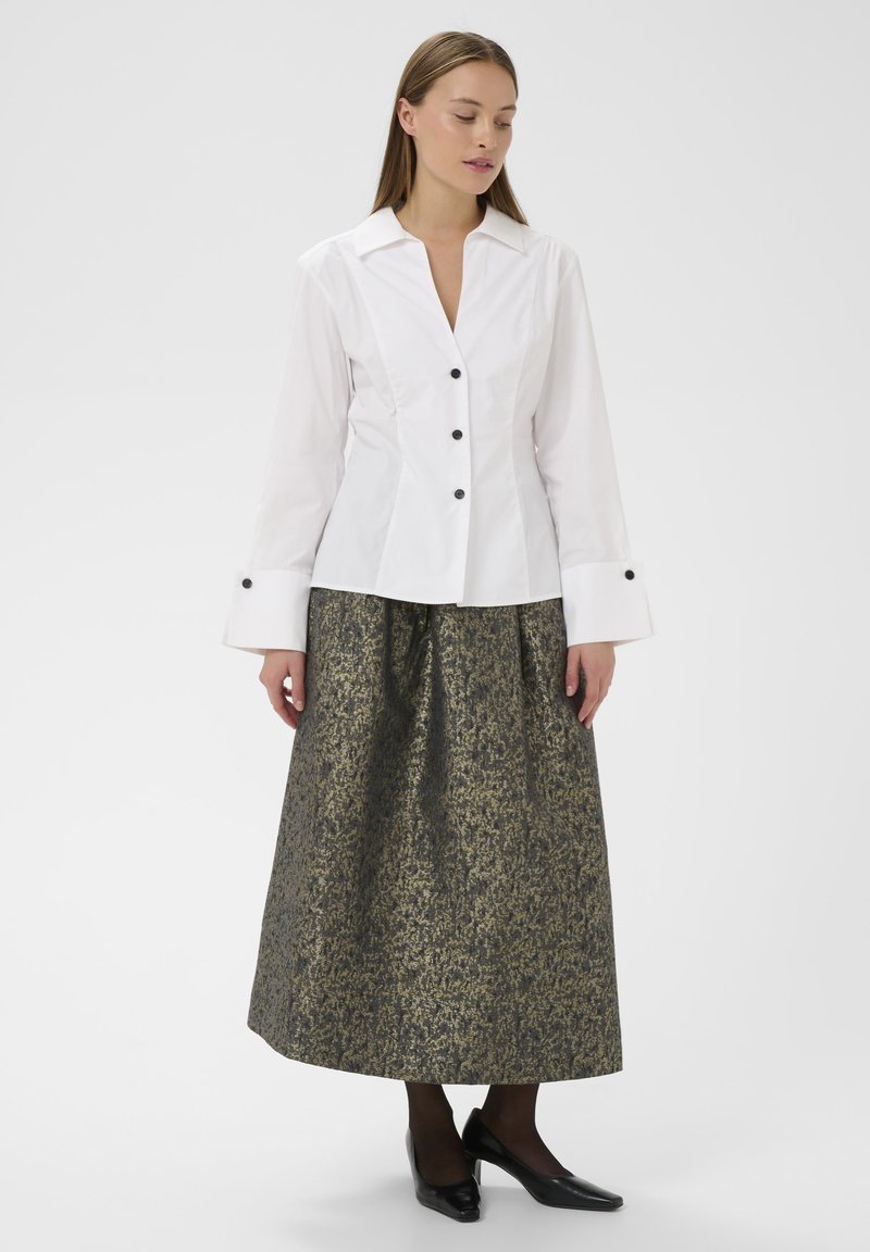 Bijela košulja s dugim rukavima, uparena s suknjom do koljena sa zlatnim i crnim uzorkom. Crne štikle upotpunjuju odjevnu kombinaciju.