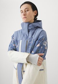 Jakna s blokovima boja u plavim i krem nijansama, cvjetni uzorak na rukavima, visoki ovratnik i rupice za palceve u manžetama za dodatnu zaštitu.
