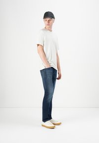 Young man wearing a black cap, off-white T-shirt, blue jeans, and white sneakers stands looking to the side with one hand in pocket.