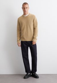 Beige Rippstrickpullover mit langen Ärmeln, kombiniert mit dunkelblauen Jeans und schwarzen Schuhen. Der Pullover hat einen runden Halsausschnitt und eine strukturierte Stoffoberfläche.