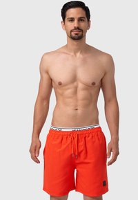 Shorts de bain orange vif avec une taille élastique et un cordon noir. Présente un petit patch logo noir sur le côté gauche.