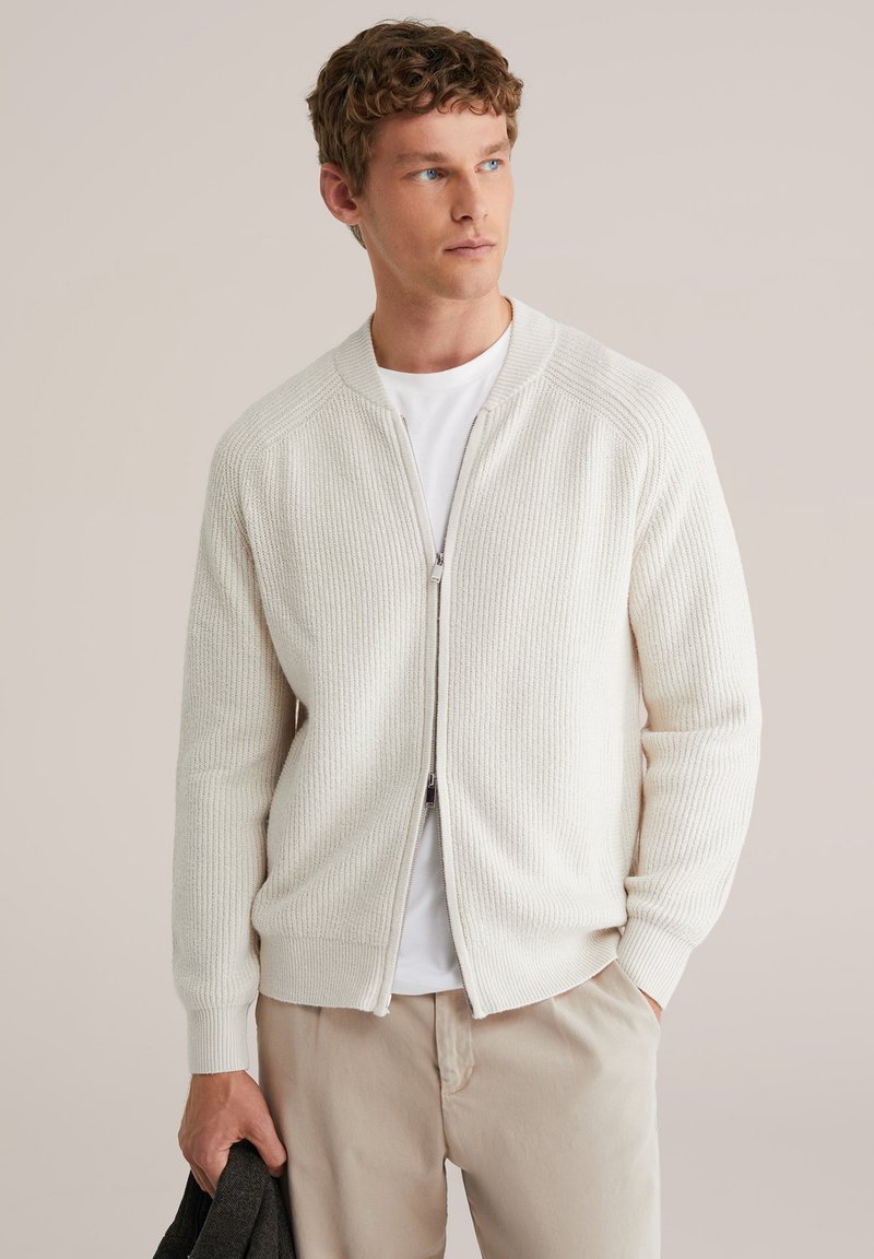 Kreemikas soonikkoes lukuga kampsun, millel on kehale mugav lõige, pikad varrukad ja selle all valge ümara kaelusega t-särk. Helebeežid püksid ja tume jakk.