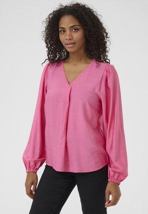 Blouse rose avec un décolleté en V, des manches bouffantes froncées et une coupe loose. Le tissu semble lisse avec un léger éclat. Associée à un pantalon noir.