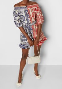 Vestido off-the-shoulder con patrones florales en azul, rojo y crema, cintura ceñida con cinturón, mangas cortas, dobladillo con volantes, combinado con un bolso blanco y tacones.