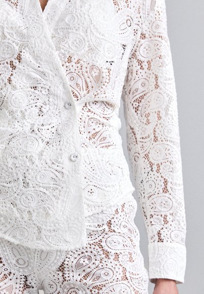 Ensemble en dentelle blanche avec des motifs en tourbillon complexes, chemise à boutons avec des manches longues, présentant une texture transparente et un aspect superposé.
