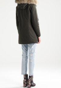 Parka vert foncé avec une capuche bordée de fourrure, associée à un jean bleu clair déchiré et des bottines noires à motif floral. Fond uni.