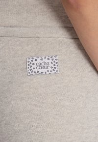 Grijze gebreide stof met een vage glans, voorzien van een wit label met zwarte luipaardvlekken en de tekst "cache coeur" duidelijk weergegeven.